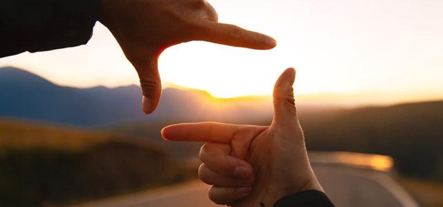 Two hands forming a rectangular frame around a sunset over a winding road and mountains.